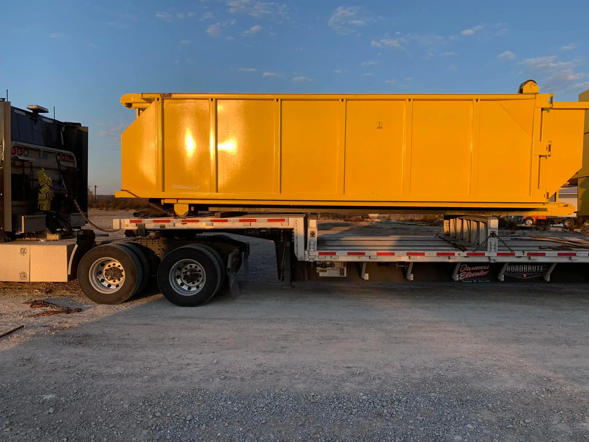 Bright yellow dumpster placed on a flatbed trailer at an open lot during sunset